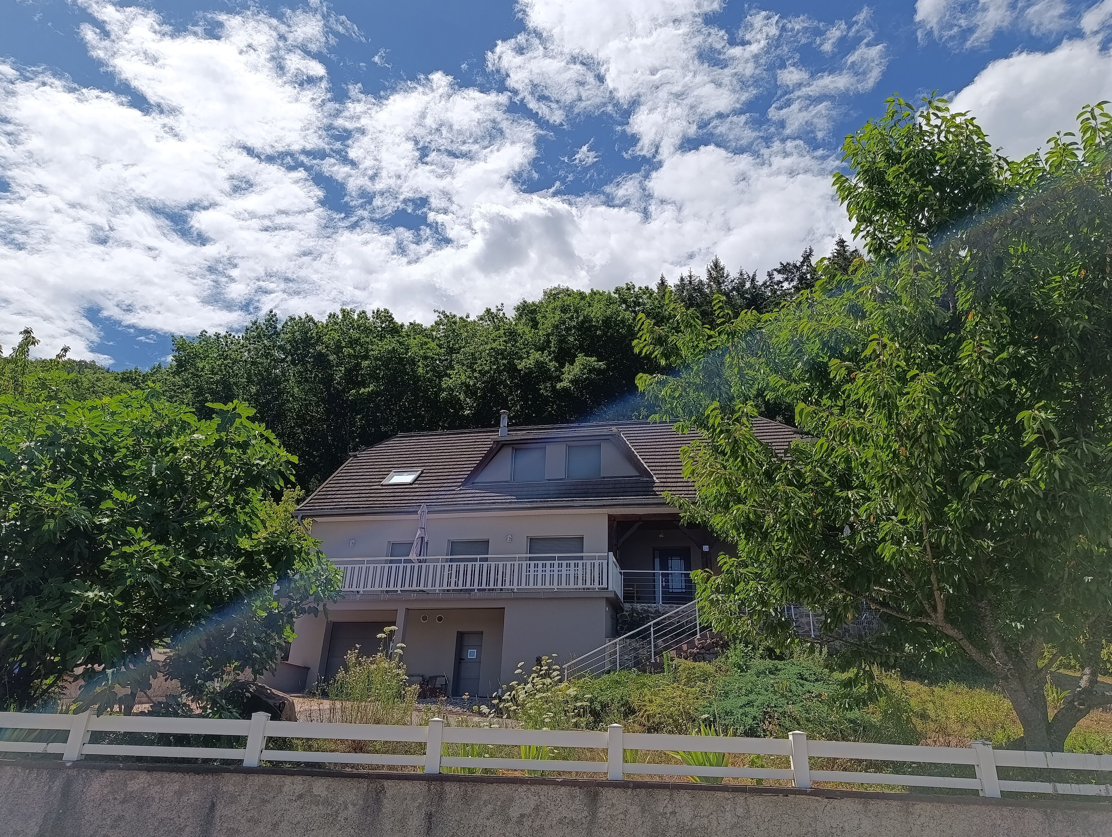 Una casa a due piani con un balcone, immersa nel verde e circondata da un recinto bianco.