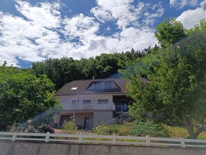 Ein zweistöckiges Haus mit Balkon, umgeben von üppiger Vegetation und einem weißen Zaun.