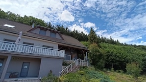 A two-story building with a balcony, a staircase leading to a covered area, and a surrounding landscape with trees and a hill.