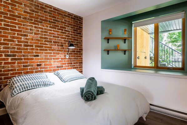 Ein Schlafzimmer mit Backsteinwand, einem Bett mit weißer Bettwäsche, einem zusammengerollten Handtuch und einem Fenster mit Blick auf eine Treppe und Grünflächen.