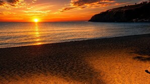 En strand med solnedgang, roligt hav og en klippe i baggrunden.