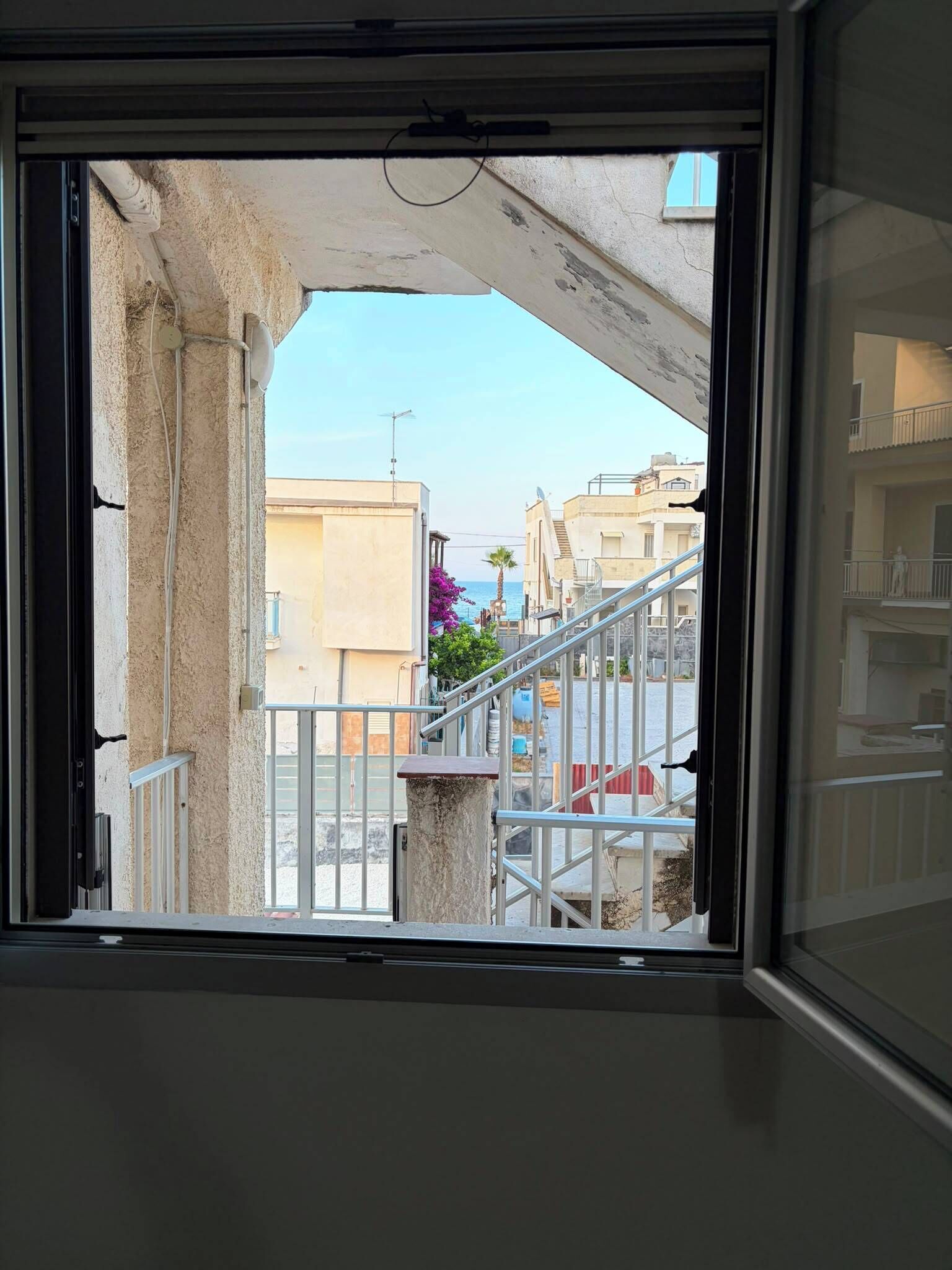 Vista a través de una ventana, mostrando una estructura de concreto por encima, una escalera de metal y edificios al fondo.