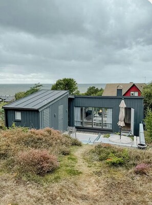 A modern two-story building with a large glass door entrance, a patio with outdoor furniture, and a view of the sea. - Architect-designed cottage where sea meets forest (Haverdal)