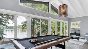 Mesa de billar con vista a un lago, ventanas amplias y una lámpara colgante.