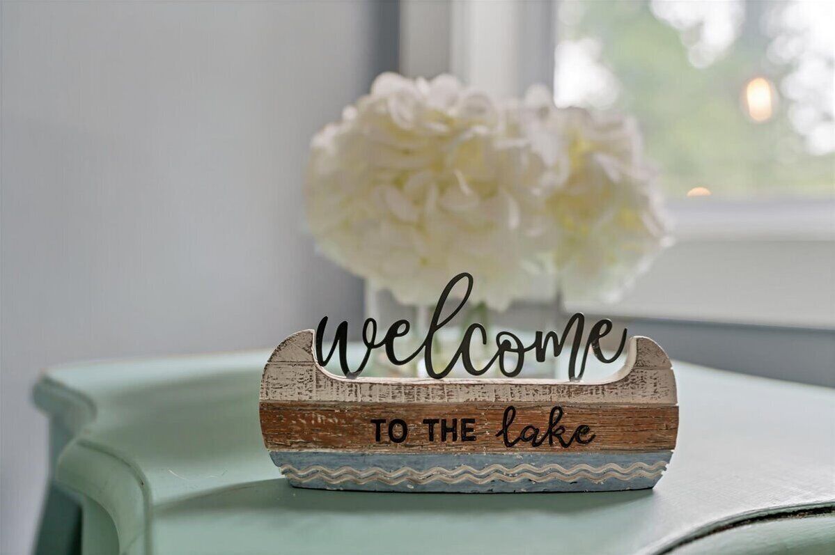 A rustic welcome sign with 'welcome to the lake' on a wooden base, placed on a table with a vase of white flowers in the background.