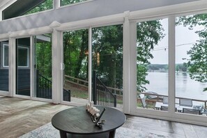 A spacious room with large windows overlooking a body of water, a round table with a decorative item, and a view of a deck area. - Relaxing Badin Lake Waterfront l Private Pier,Hot Tub,Pool Table-Dock Your Boat! (New London)