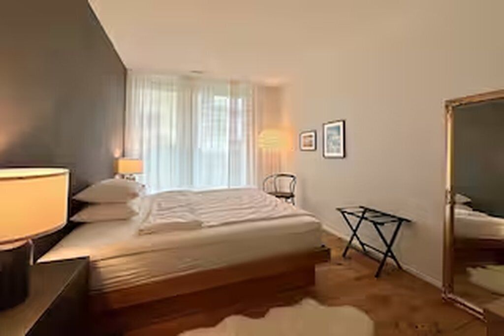 A bedroom with a bed, a bedside lamp, a window with curtains, a small table, and a mirror.