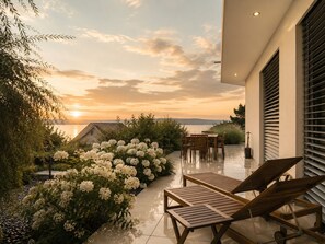 Un patio con vistas a la puesta de sol sobre un cuerpo de agua, sillones de madera para relajarse y un jardín con flores blancas.
