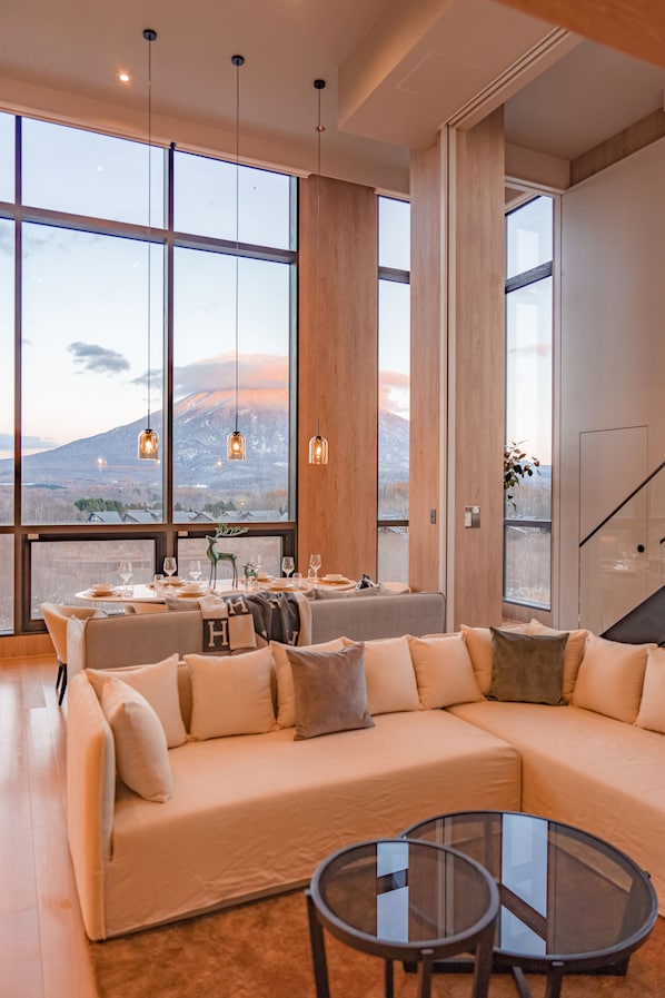 A modern living room with a large sectional sofa, a glass coffee table, and a dining area with a view of a mountainous landscape. - The 1898 Niseko (Kutchan)