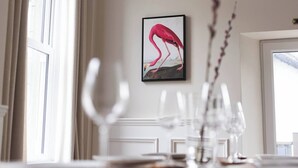 Uma área de refeições com uma mesa posta para dois, uma obra de arte emoldurada de um flamingo na parede e uma janela com cortinas.