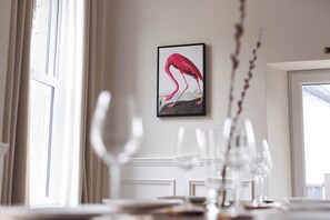 A dining area with a table set for two, a framed artwork of a flamingo on the wall, and a window with curtains.