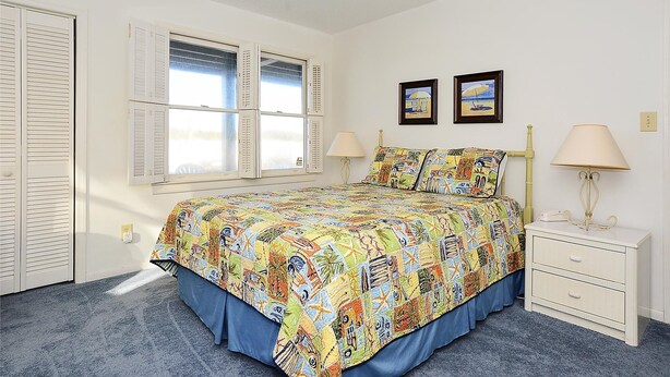 A bedroom with a bed, two bedside lamps, a nightstand, a window with shutters, and two framed pictures on the wall. - Shore to Please: Oceanfront, Pool, Classic Beach Charm (Murrells Inlet)