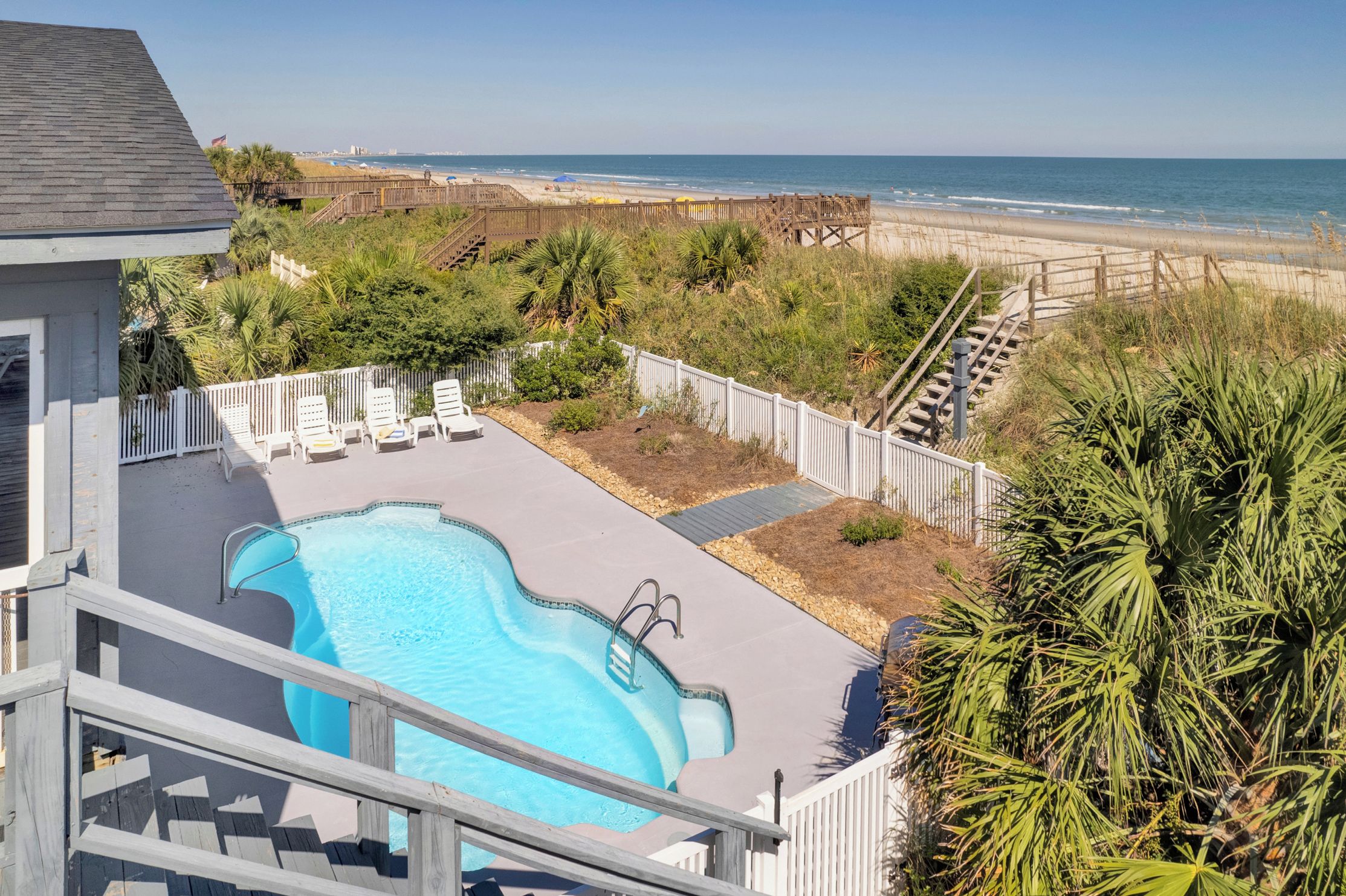 A swimming pool with a wooden deck, surrounded by a white fence and palm trees, overlooking a beach.