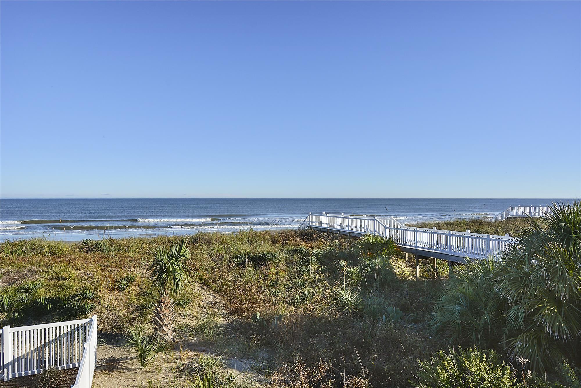A coastal view with a white wooden walkway leading to the beach, surrounded by dense vegetation.