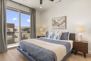 A bedroom with a large bed, a nightstand, a lamp, a painting, and a view of a building through a window. - Splash And Swing 6 Bedroom Townhouse (St. George)