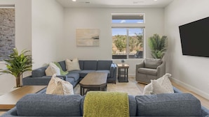 A modern living room with a gray sectional sofa, a wooden coffee table, a gray armchair, a small side table, and a large window with a view of the outdoors. - Splash And Swing 6 Bedroom Townhouse (St. George)