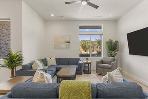 Ein modernes Wohnzimmer mit einem grauen Ecksofa, einem Holztisch, einem grauen Sessel, einem kleinen Beistelltisch und einem großen Fenster mit Blick ins Freie.
