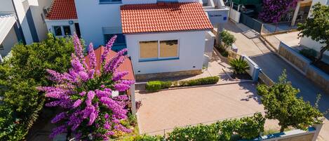 A two-story building with a red-tiled roof, a small balcony, and a garden with purple flowers.