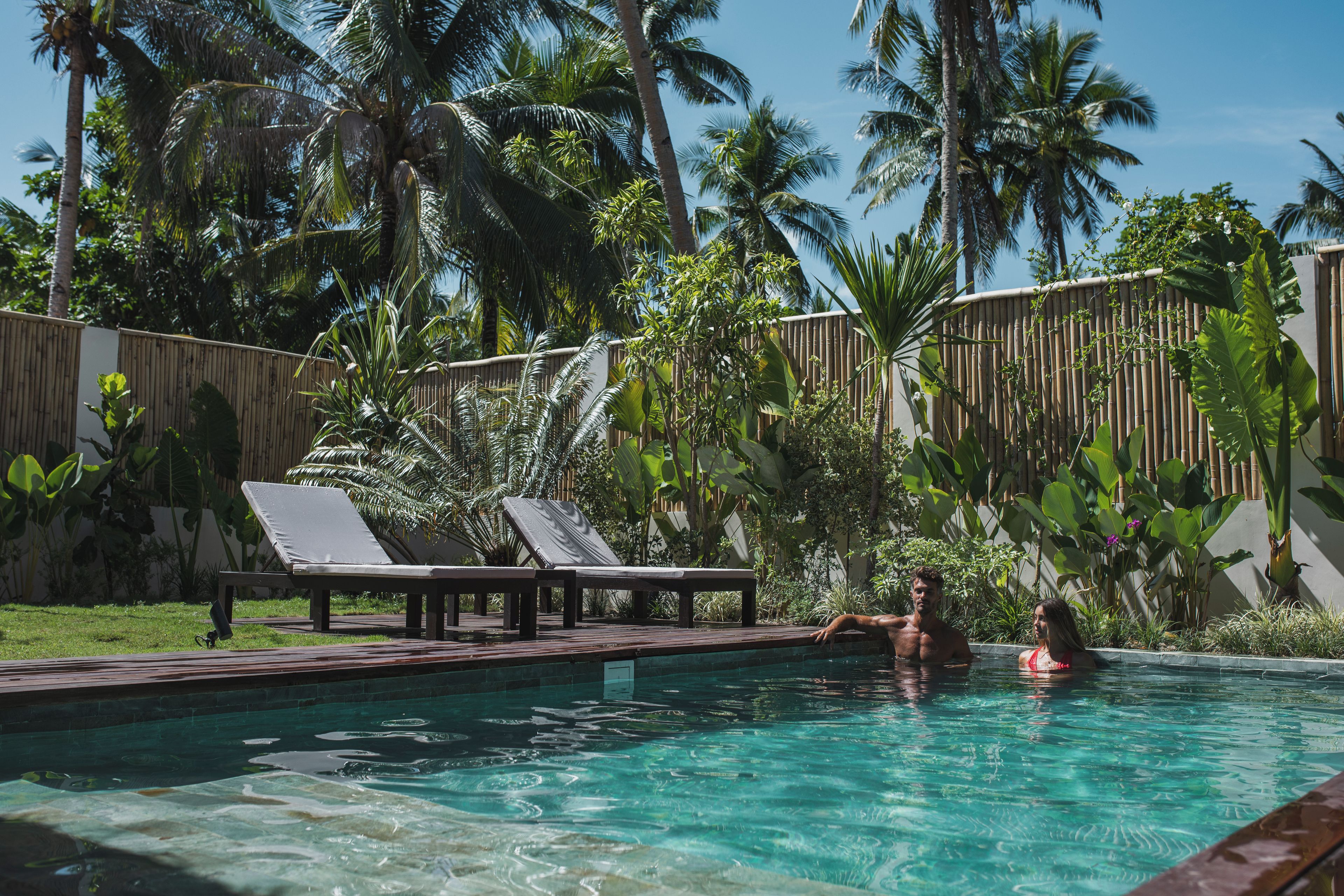 A swimming pool with lounge chairs, surrounded by tropical vegetation and a bamboo fence.