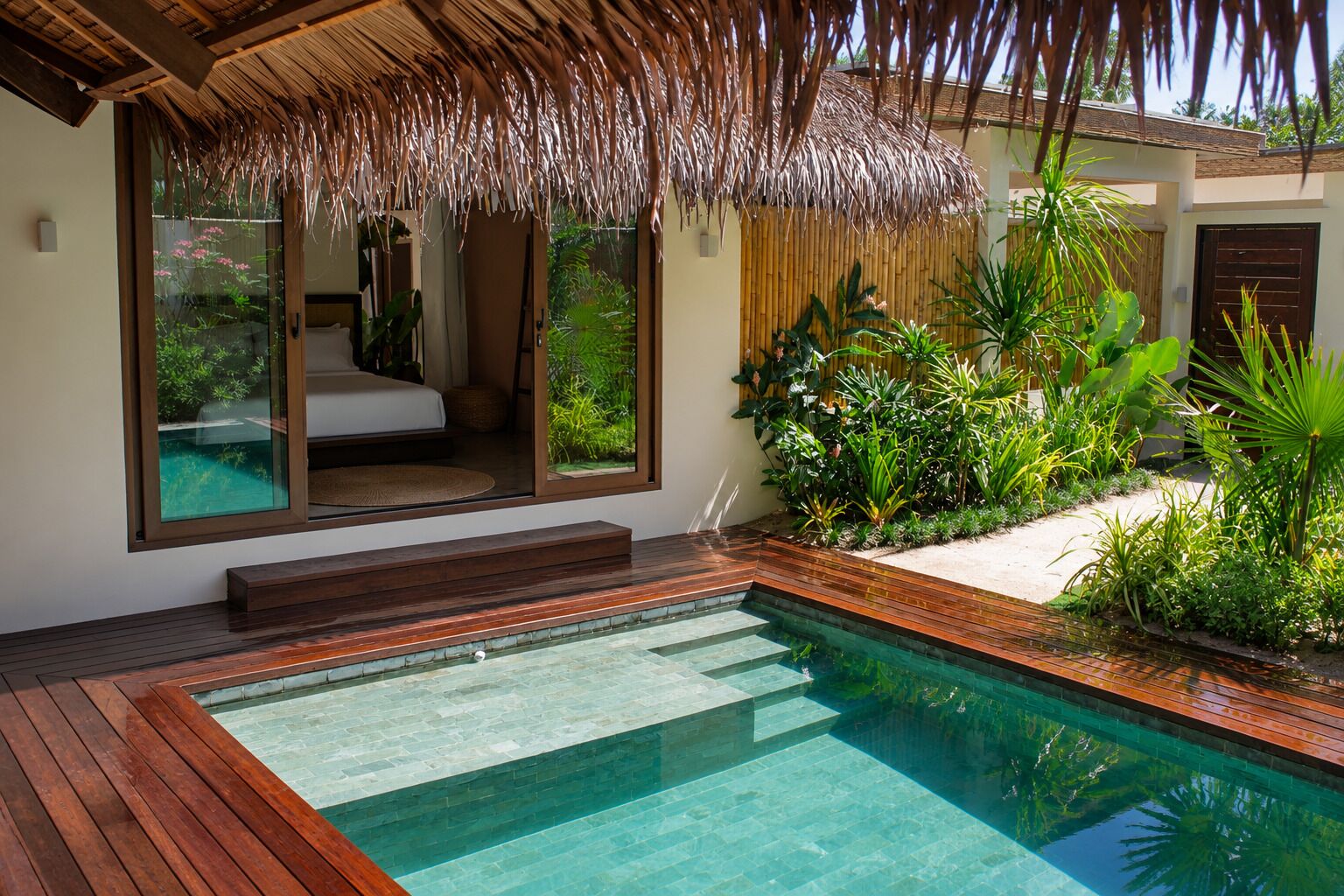 A modern outdoor pool area with a wooden deck, a glass-enclosed bedroom, and lush tropical plants.