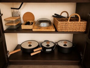 Una repisa de cocina con un rallador, una tabla de cortar de madera, un colador metálico, una cesta tejida y tres ollas con asas de madera.