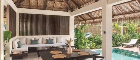 A modern indoor pool area with a thatched roof, a dining table with chairs, and a seating area with a sofa and cushions.