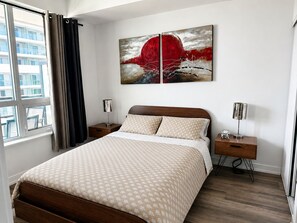 A modern hotel room with a large bed, a nightstand, a lamp, and a painting on the wall.