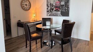 A dining area with a round glass table, four chairs, a wall clock, and a black and white photograph of a red double-decker bus.