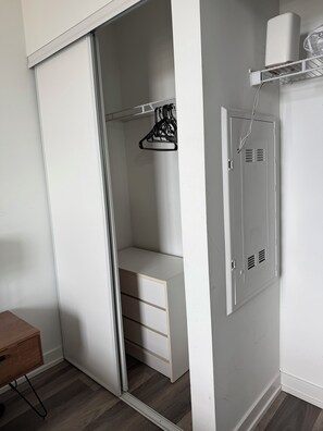 Un armari blanc amb porta corredissa, una prateleira amb roba penjada i una presa elèctrica a la paret.