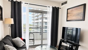 A modern living room with a glass table, a sofa, a TV, and a balcony with a view of a building.