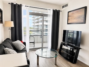 A modern living room with a glass table, a sofa, a TV, and a balcony with a view of a building.