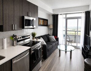 Una cocina moderna con estufa, horno y microondas. Hay una zona de comedor con mesa de vidrio y sofá. La habitación tiene un ventanal con cortinas.