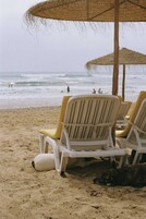 Beach chairs under a straw umbrella on a sandy beach with the ocean in the background.
