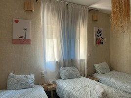 Una habitación con tres camas, una ventana con cortinas traslúcidas, un cuadro con motivos del desierto y una planta colgante.
