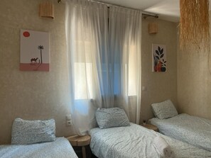 A room with three beds, a window with sheer curtains, a wall with a desert-themed artwork, and a hanging plant.