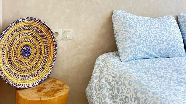 A bedroom with a bed, a wall-mounted light, a patterned rug, and a small wooden stool.