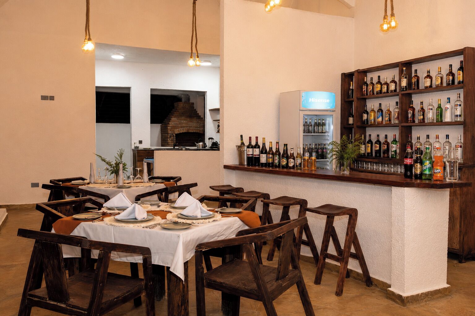 A restaurant with wooden tables and chairs, a bar area with a variety of bottles, and a brick oven in the background.