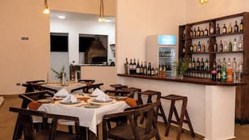 A restaurant with wooden tables and chairs, a bar area with a variety of bottles, and a brick oven in the background.