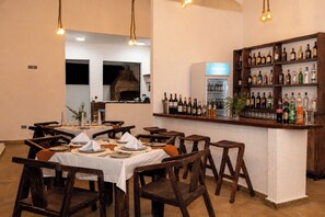 A restaurant with wooden tables and chairs, a bar area with a variety of bottles, and a brick oven in the background. - Olarashii River Lodge (Arusha)