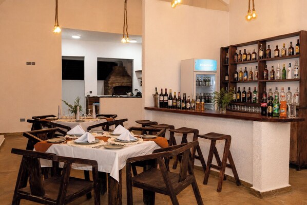 Un restaurant avec des tables et des chaises en bois, un bar bien garni de bouteilles variées, et un four en briques à l’arrière-plan.