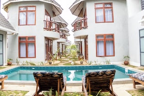 Zona de piscina con sillas de descanso, piscina y un edificio con varios balcones.