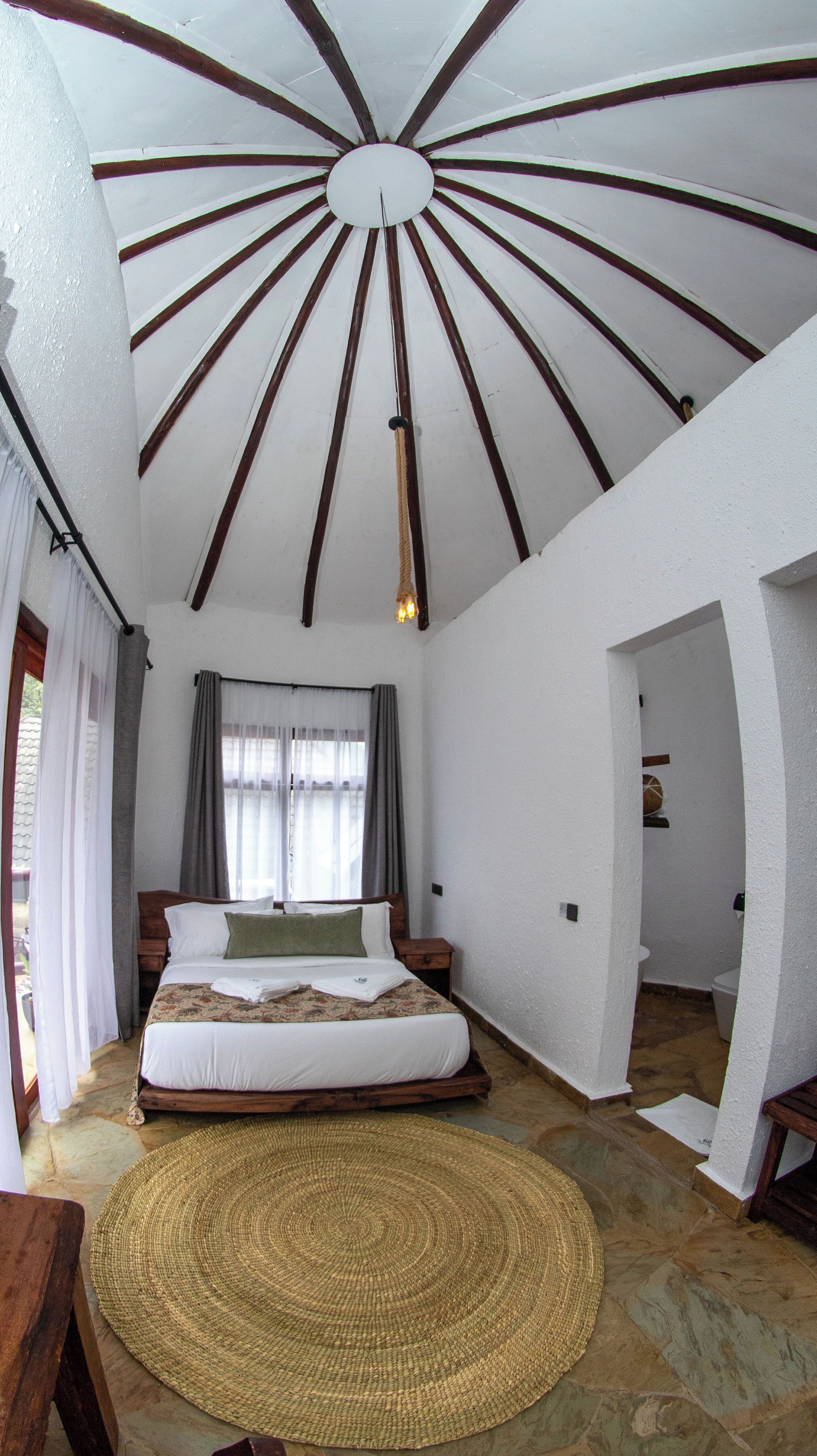 Um quarto com cama de madeira, tapete tecido, teto com vigas de madeira e janela com cortinas.