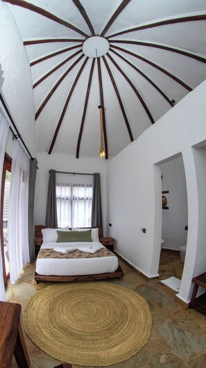 A bedroom with a wooden bed, a woven rug, a ceiling with wooden beams, and a window with curtains. - Olarashii River Lodge (Arusha)