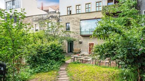 A garden pathway leading to a seating area with a bench and table, surrounded by lush greenery and a building with stone walls and windows.