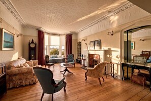 A room with a fireplace, wooden flooring, and classic furniture including a sofa, armchairs, and a table.