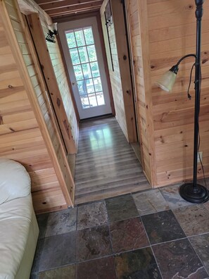 A wooden interior hallway with a door leading outside, a floor lamp, and a bed.