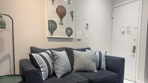 A dark blue sofa with striped pillows, a glass side table, a wall-mounted light fixture, and a door with a white frame. - Discover the Comfort of Columbia University Area-2 (New York)