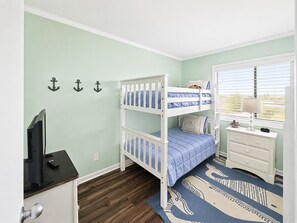 A bunk bed with blue bedding, a desk with a computer, and a window with blinds. - Dunescape Villas 108 (Atlantic Beach)