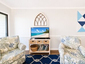 A living room with a television on a wooden stand, two patterned armchairs, a woven basket, and a geometric wall art piece. - Dunescape Villas 108 (Atlantic Beach)