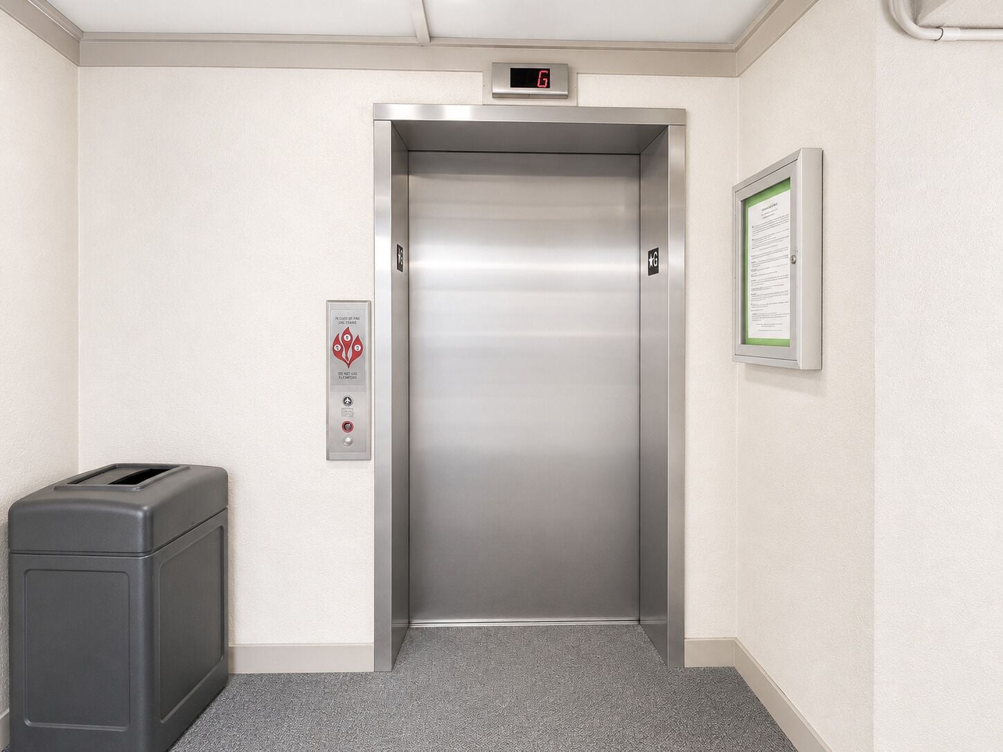 An elevator with a digital display, a fire alarm, and a trash bin.